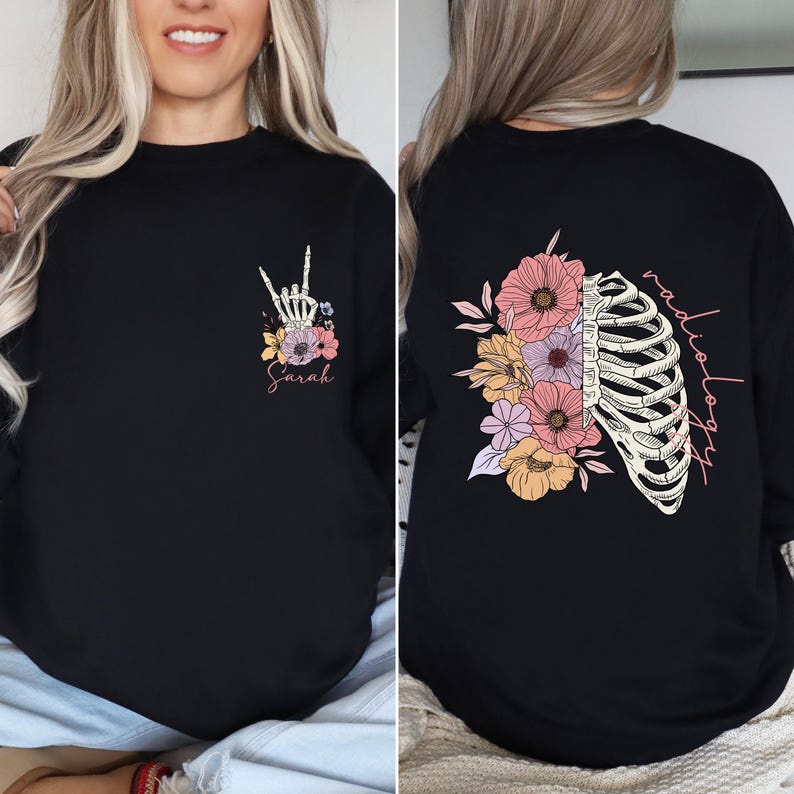 Pode incluir: Camiseta preta de manga comprida com um design floral e uma caixa tor&aacute;cica de esqueleto nas costas. O texto "radiologia" est&aacute; escrito verticalmente do lado direito da caixa tor&aacute;cica. A frente da camiseta tem uma m&atilde;o de esqueleto fazendo o sinal de rock and roll com flores ao redor. O texto "Sarah" est&aacute; escrito abaixo da m&atilde;o.