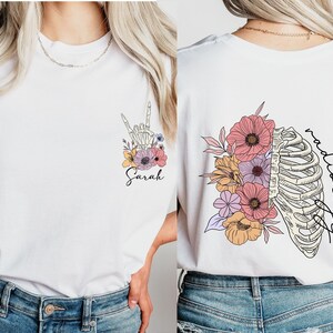 Pode incluir: Camiseta branca com um design floral e uma m&atilde;o de esqueleto fazendo o sinal de linguagem de sinais 'Eu te amo'. O texto 'Sarah' est&aacute; abaixo do design. A parte de tr&aacute;s da camiseta tem um design floral com uma caixa tor&aacute;cica e o texto 'radiologia'.