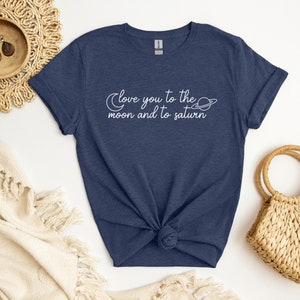 Può includere: Una maglietta blu navy con una grafica bianca che dice "Love you to the moon and to saturn" con una luna crescente e un design ad anello di pianeta.