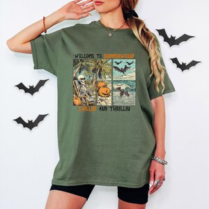 May include: Olive green t-shirt with a graphic design featuring a skeleton relaxing on a beach, pumpkins, bats, and the text "WELCOME TO SUMMERWEEN, CHILLIN AND THRILLIN". The shirt is worn by a person.
