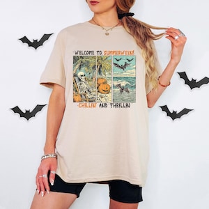 May include: Beige t-shirt with a graphic that reads "WELCOME TO SUMMERWEEN! CHILLIN AND THRILLIN". The graphic features a skeleton on a beach, pumpkins, bats, and ocean waves. Black shorts and bat decorations are also visible.