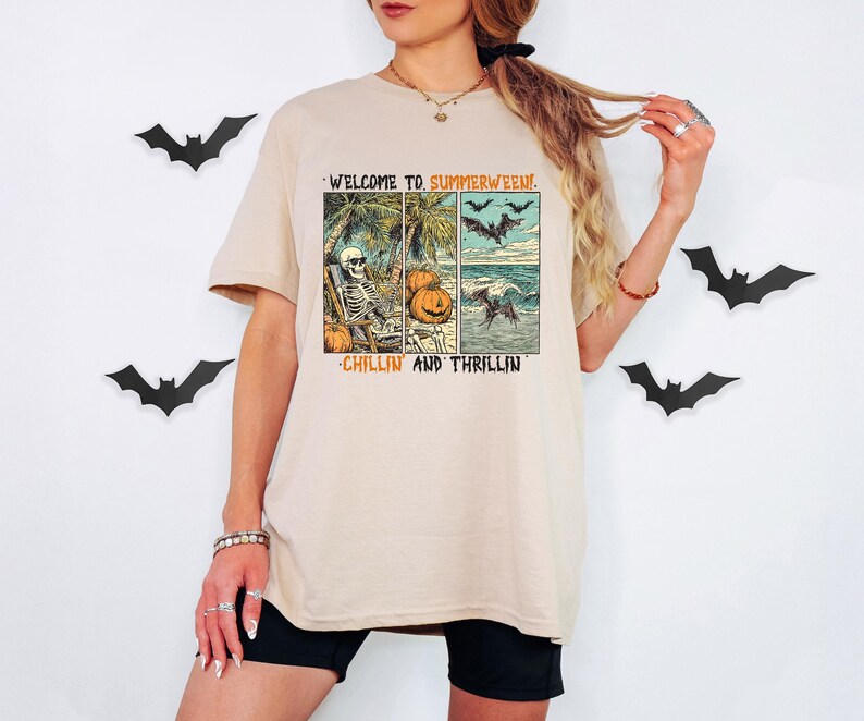 May include: Beige t-shirt with a graphic that reads "WELCOME TO SUMMERWEEN! CHILLIN AND THRILLIN". The graphic features a skeleton on a beach, pumpkins, bats, and ocean waves. Black shorts and bat decorations are also visible.