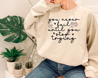 May include: A beige sweatshirt with the text "you never fail until you stop trying" in black script with small pink flowers and hearts.
