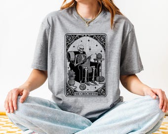 May include: A gray t-shirt with a black and white illustration of a skeleton crocheting in a chair. The illustration is framed with a decorative border and the text "The Crocheter" is below the image.