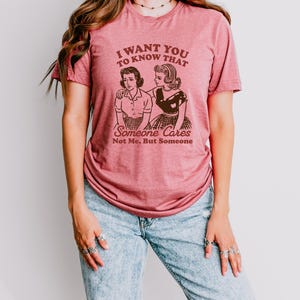 Op de afbeelding: Een roze T-shirt met de tekst "Ik wil dat je weet dat iemand geeft, niet ik, maar iemand" en een retro-illustratie van twee vrouwen.