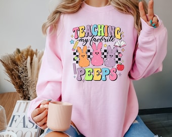 May include: Pink sweatshirt with the text "Teaching my favorite peeps" in rainbow colors and a graphic of three pastel-colored Peeps chicks.