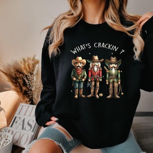 May include: Black sweatshirt featuring three cowboy nutcrackers with hats, mustaches, and cowboy attire. The text "WHAT'S CRACKIN ?" is arched above the nutcrackers. The sweatshirt is a casual, festive garment.