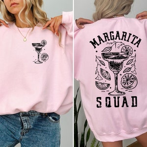 Peut inclure: Sweat-shirt rose avec un graphique noir d'un verre &agrave; margarita avec des tranches de citron vert et le texte "Margarita Squad" au dos.