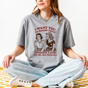 Op de afbeelding: Een grijs T-shirt met een vintage-stijl illustratie van twee vrouwen. De tekst op het shirt luidt "Ik wil dat je weet dat iemand geeft, niet ik, maar iemand."