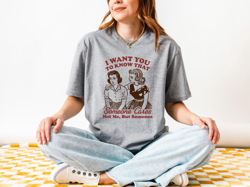 Op de afbeelding: Een grijs T-shirt met een vintage-stijl illustratie van twee vrouwen. De tekst op het shirt luidt "Ik wil dat je weet dat iemand geeft, niet ik, maar iemand."
