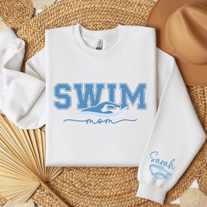 Pu&ograve; includere: Felpa bianca con la scritta "SWIM" in lettere blu, una grafica a onda e la parola "mom" in corsivo. La manica ha il nome "Sarah" e un paio di occhialini blu. La felpa &egrave; su una superficie tessuta.