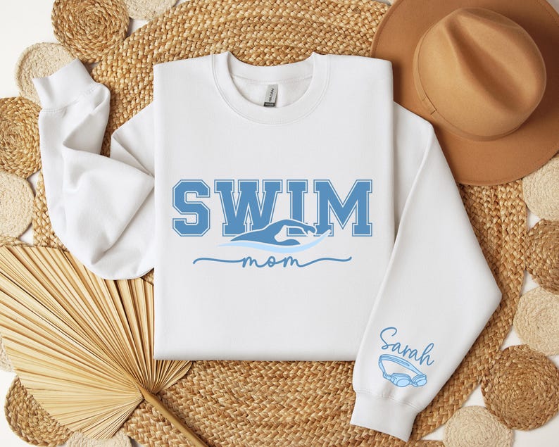 Pu&ograve; includere: Felpa bianca con la scritta "SWIM" in lettere blu, una grafica a onda e la parola "mom" in corsivo. La manica ha il nome "Sarah" e un paio di occhialini blu. La felpa &egrave; su una superficie tessuta.