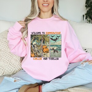 May include: Pink sweatshirt with a graphic that reads "WELCOME TO. SUMMERWEEN!" and "CHILLIN AND THRILLIN". The graphic features a skeleton relaxing on a beach, pumpkins, bats, and a wave with a bat.