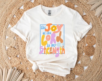 May include: A white t-shirt with a colorful graphic design that says "the joy of the lord is my strength". The graphic features a flower, a cross, and a heart.