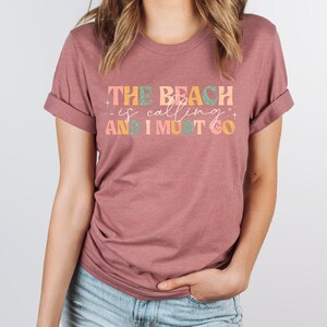 May include: A mauve t-shirt with the text "THE BEACH is calling AND I MUST GO" in colorful letters.