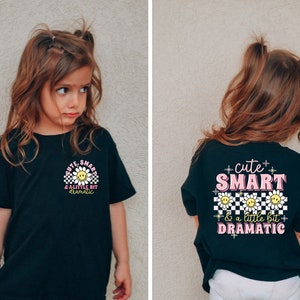 May include: Black t-shirt with a pink and white graphic that says "cute smart & a little bit dramatic" with a smiley face and checkered pattern.