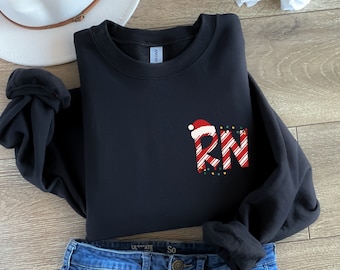 May include: Black sweatshirt with a Christmas-themed RN design. The design features the letters "RN" in red and white candy cane stripes, a Santa hat, and string lights. The sweatshirt is folded with a white hat and jeans in the background.
