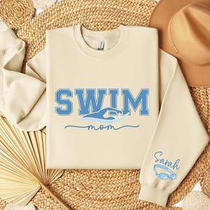 Pu&ograve; includere: Felpa color crema con la scritta "SWIM" in lettere blu, una grafica di nuotatore e la parola "mom". La manica destra presenta il nome "Sarah" e un paio di occhialini blu. La felpa &egrave; esposta su una superficie tessuta.