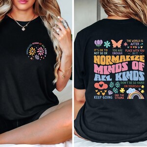 May include: Black t-shirt with a colorful graphic print that says "Normalize Minds of All Kinds". The graphic includes flowers, hearts, a rainbow, and the text "It's ok to not be ok", "You are enough", "The world is after... Place with you in it", "Don't be so hard on yourself", "Keep going", and "End the stigma".