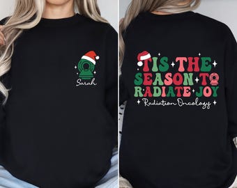 May include: Black sweatshirt with a festive design. The front displays a medical imaging machine with a Santa hat, the name "Sarah", and stars. The back says "Tis the Season to Radiate Joy" with "Radiation Oncology" below.