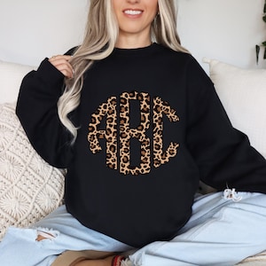 May include: Black sweatshirt featuring a leopard print monogram design. The monogram includes the letters "ABC" in a stylish, rounded font. The leopard print pattern has brown and tan spots on a light brown background. The sweatshirt is a casual, comfortable style.