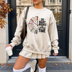 May include: A cream-colored sweatshirt with the text "SO MANY BOOKS SO LITTLE TIME" in black. The design features a clock face made of colorful books. The sweatshirt is paired with a pleated skirt and white boots.