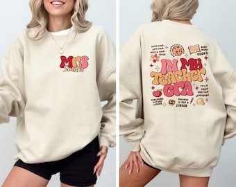 May include: A beige sweatshirt with a retro-style graphic on the front that says "Mrs. Smith" and a retro-style graphic on the back that says "In My Teacher Era" with various teacher-themed icons.