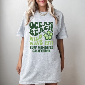 May include: Gray t-shirt with a green graphic print that says "Ocean Beach Wild Wave 1971 Surf Memories California" with a green hibiscus flower.