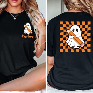 May include: Black t-shirt featuring a cartoon ghost holding a pencil. The front of the shirt has the text "Mrs. Grey" in orange. The back of the shirt has the ghost and pencil design on a checkered orange and black background.