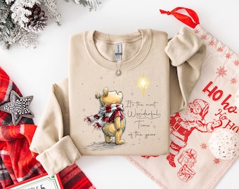 Op de afbeelding: Een beige sweatshirt met een afbeelding van een beer met een sjaal en de tekst "It's the most Wonderful Time of the Year". Een kerstcadeautas en een rood-zwarte geruite sjaal zijn ook zichtbaar.