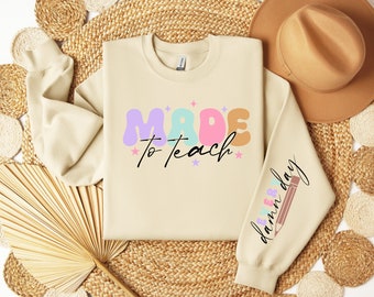 Może przedstawiać: Beżowa bluza z napisem "MADE to teach" w kolorowych literach i ołówkiem z napisem "every day" na rękawie.