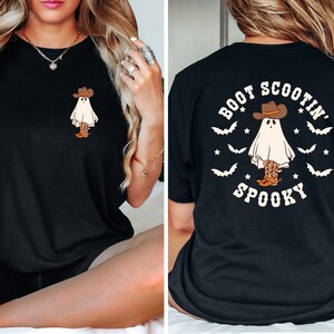 May include: Black t-shirt featuring a cartoon ghost wearing a cowboy hat and boots. The front of the shirt has a small graphic of the ghost, while the back displays the text "Boot Scootin' Spooky" in a circular design with bats and stars.