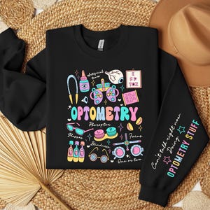 May include: Black sweatshirt with a colorful graphic design featuring the word "OPTOMETRY" and various eye-related illustrations, including glasses, an eye chart, and eye drops. The right sleeve has the text "Can't talk right now Doing OPTOMETRY STUFF" in a colorful font.