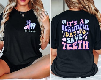 Może przedstawiać: Czarny T-shirt z wzorem o tematyce dentystycznej. Przód koszulki przedstawia ząb z szczoteczką do zębów, nicią dentystyczną i pastą do zębów. Tył koszulki głosi "It's a beautiful day to save teeth" kolorowymi literami w stylu retro.