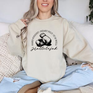 May include: Beige sweatshirt with a graphic of boxing gloves and the words "Hard Fought, Heart Felt, It Is Well, Hallelujah" in black. The sweatshirt is a neutral color and has a crew neck.