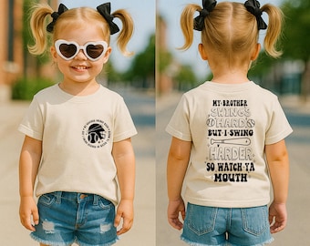 May include: A young girl wearing a beige t-shirt with a baseball graphic on the front and the text "My Brother Swings Hard But I Swing Harder So Watch Ya Mouth" on the back. She is wearing denim shorts and has her hair in pigtails.