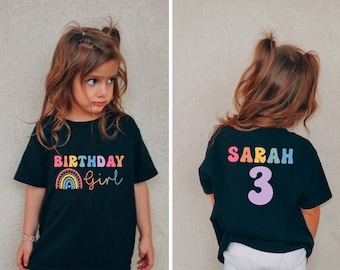 以下が含まれることがあります： 虹と「Birthday Girl」の文字が書かれた黒いTシャツ。後ろには「SARAH」と「3」の文字が書かれている。