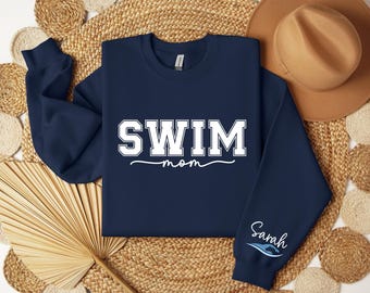 May include: Navy blue sweatshirt with white text that reads "SWIM mom" and a white wave design on the left sleeve with the text "Sarah".