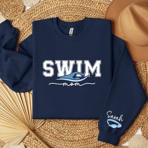 Pu&ograve; includere: Felpa blu navy con la scritta "SWIM" in lettere bianche a blocchi sopra un'onda blu e la parola "mom" in corsivo. La manica presenta il nome "Sarah" e un paio di occhialini blu.