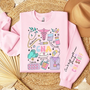 May include: Pink sweatshirt with a colorful graphic design of medical tools and the text "CNA" in large letters. The sleeve has the text "Can't talk right now Doing CNA STUFF".