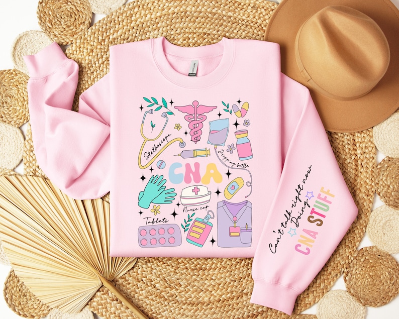 May include: Pink sweatshirt with a colorful graphic design of medical tools and the text "CNA" in large letters. The sleeve has the text "Can't talk right now Doing CNA STUFF".