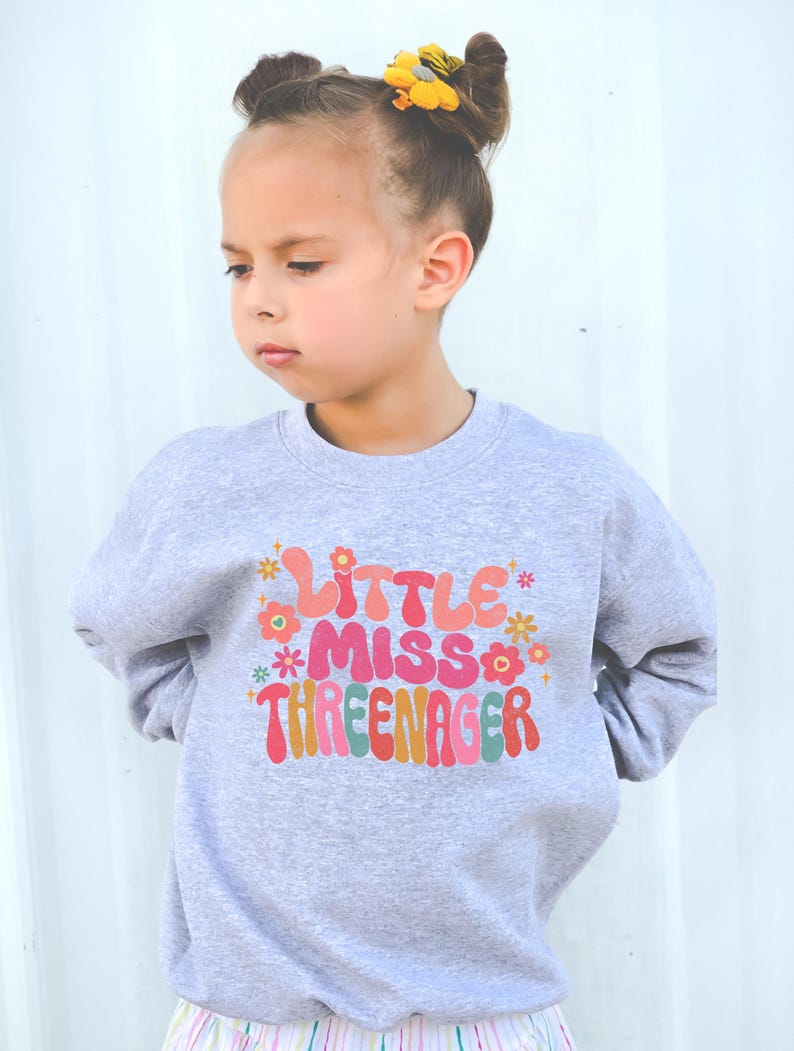 May include: A gray sweatshirt with the text "Little Miss Three-nager" in colorful, retro-style lettering. The text is surrounded by small, colorful flowers.