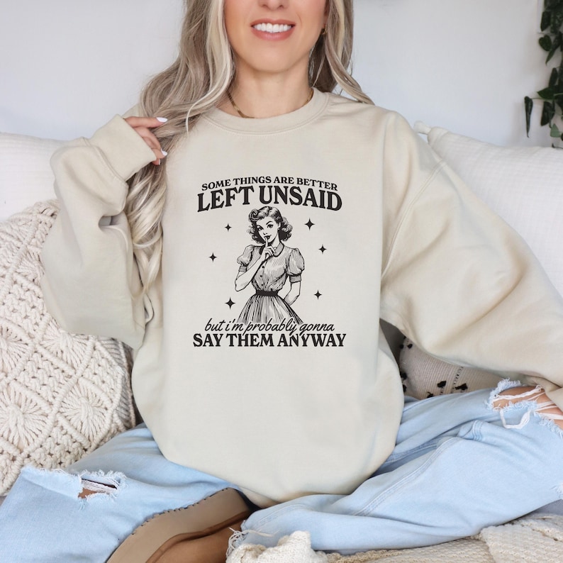 May include: A beige sweatshirt with a retro-style illustration of a woman with her finger to her lips. The text on the sweatshirt reads "Some things are better left unsaid, but I'm probably gonna say them anyway."
