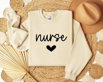 Może przedstawiać: Beżowa bluza z nadrukiem słowa "nurse" i czarnego serca z przodu.
