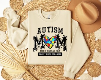 May include: A beige sweatshirt with the text "AUTISM MOM" in black block letters. A heart-shaped puzzle piece design in the center features various colors. The text "LIKE A NORMAL MOM EXCEPT MUCH STRONGER" is below the heart.