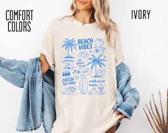 Może przedstawiać: Kremowa koszulka z niebieską grafiką o tematyce plażowej. Projekt zawiera palmy, słońce, fale oraz napisy "Beach Vibes", "Sunshine", "Catch the Wave" i "Meet me at beach."