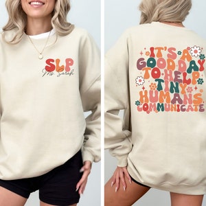 May include: A beige sweatshirt with the text "SLP Ms. Sarah" on the front and "It's a good day to help tiny humans communicate" on the back. The text is in a retro font with colorful accents.