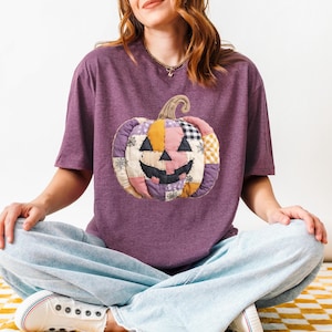 May include: A purple t-shirt featuring a patchwork pumpkin design. The pumpkin has a smiling face and is made of various fabric patterns and colors, including orange, pink, and black. The shirt is a casual style, perfect for Halloween.