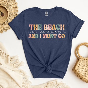 May include: A navy blue t-shirt with the text "THE BEACH is calling AND I MUST GO" in colorful, retro-style lettering.