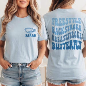 May include: Light blue t-shirt with a swimming goggles graphic and the name "SARAH" in blue. The back of the shirt features the words "FREESTYLE, BACKSTROKE, BREASTSTROKE, BUTTERFLY" in a wavy blue font. A casual, sporty design.